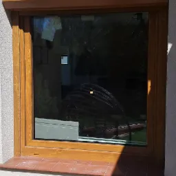 Brązowa rama okienna z zamontowaną roletą zewnętrzną, widoczna z zewnątrz budynku, z terakotowym parapetem i odbiciem w szybie.