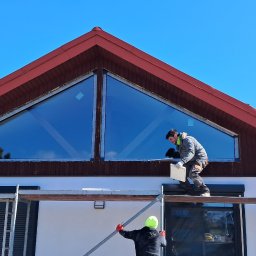 Tomasz Chałat SET - Montażysta na rusztowaniu, instalujący okno trójkątne pod dachem domu z czerwonymi elementami drewnianymi, widoczne również rolety zewnętrzne na niższych oknach.