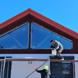 Montażysta na rusztowaniu, instalujący okno tr&oacute;jkątne pod dachem domu z czerwonymi elementami drewnianymi, widoczne r&oacute;wnież rolety zewnętrzne na niższych oknach.