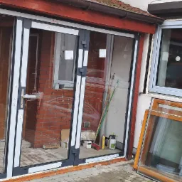 Montaż szklanych drzwi przesuwnych z szarymi ramami; obok okno z białą ramą i stos drewnianych ram okiennych; widoczne narzędzia i akcesoria montażowe.