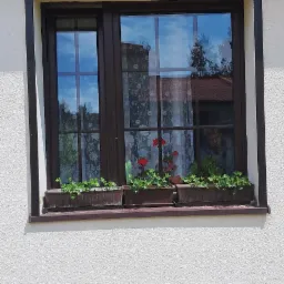 Brązowe okno PCV z czarną roletą zewnętrzną częściowo opuszczoną. Na parapecie doniczki z kwiatami. W szybie odbija się niebo i budynki.