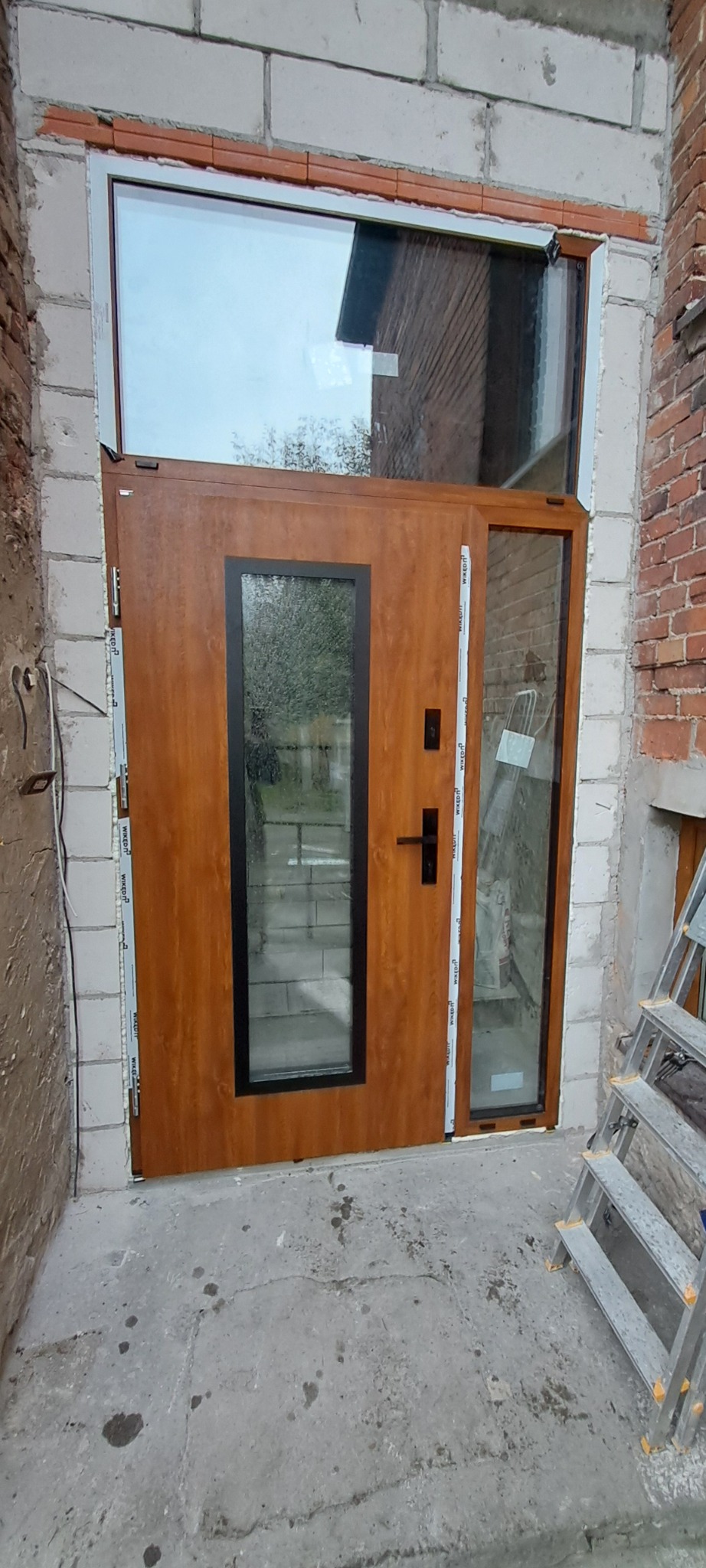 Brązowe drzwi wejściowe z szybą i górnym doświetleniem zamontowane w surowym murze z bloczków i cegieł, widoczna drabina malarska.