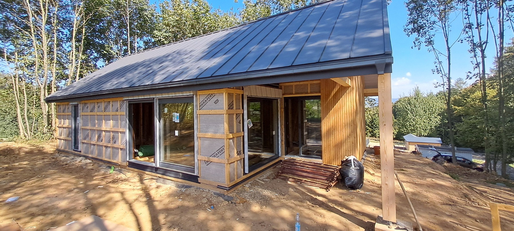 Dom w trakcie budowy z widocznymi dużymi oknami przesuwnymi, drewnianą elewacją i szarą blachą na dachu, na tle drzew