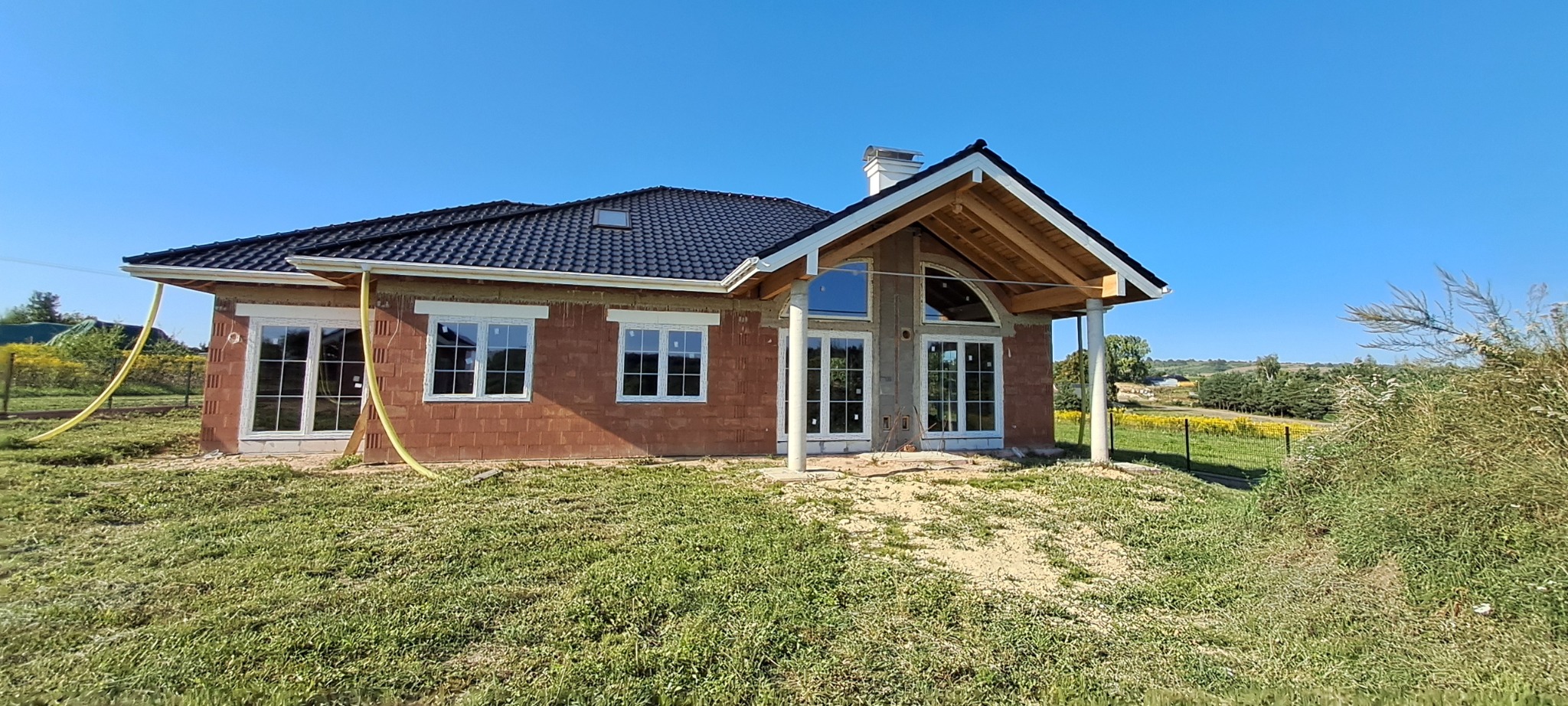 Nowo budowany dom z białymi ramami okiennymi i przeszklonymi drzwiami tarasowymi, widok na konstrukcję dachu i kolumny podtrzymujące zadaszenie, żółty wąż leżący na trawie.