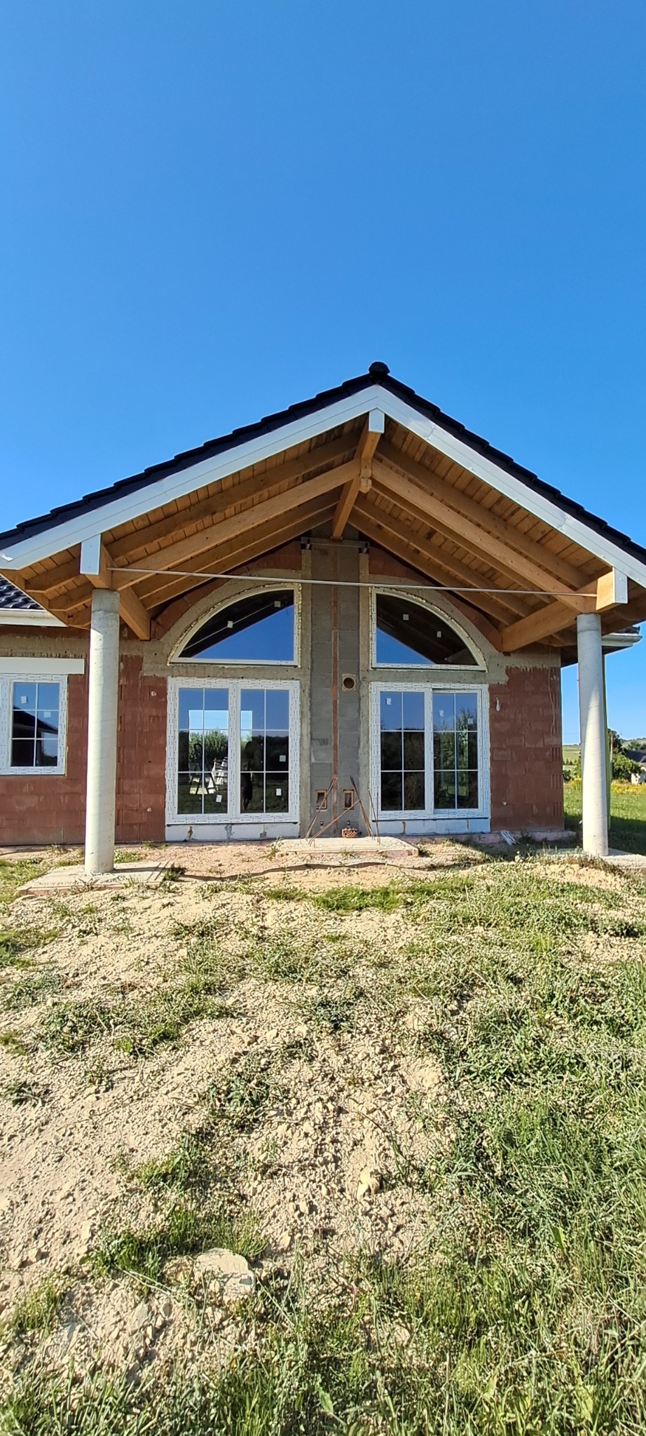 Surowy stan budowy domu jednorodzinnego z widocznymi białymi oknami o nietypowym, półokrągłym kształcie w szczycie, osadzonymi w murze z czerwonej cegły, pod drewnianym zadaszeniem wspartym...