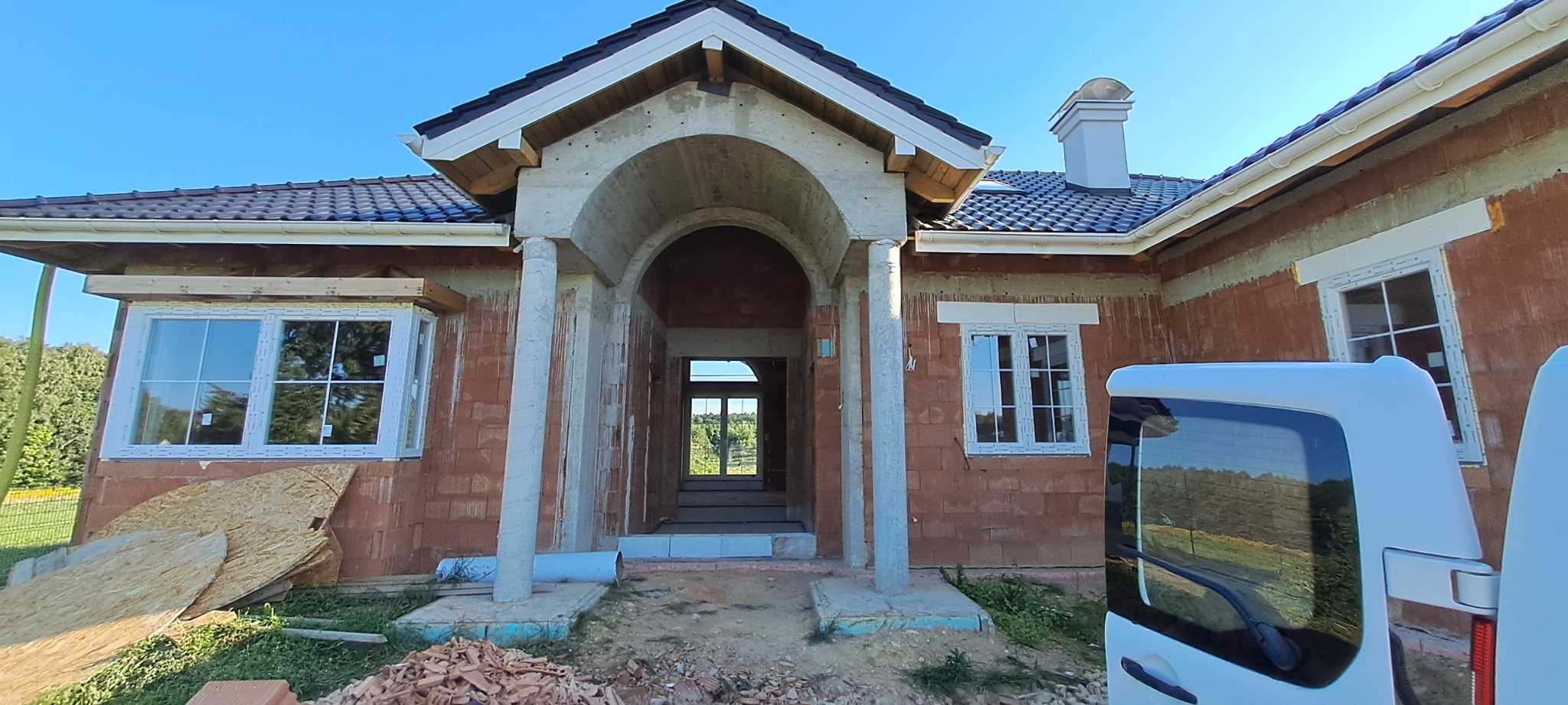 Dom w trakcie budowy z widocznymi oknami PCV, surową elewacją z czerwonej cegły i częściowo wykończonym wejściem z kolumnami.