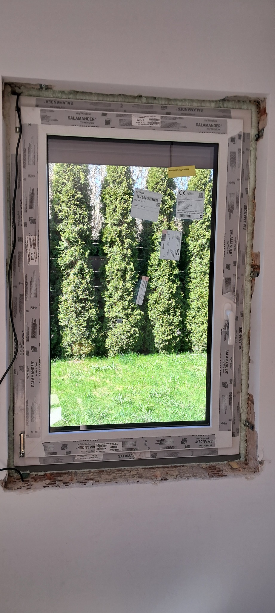 Białe okno z widokiem na ogród z tujami, oklejone folią ochronną i częściowo uszczelnione pianką montażową, widoczne etapy montażu.
