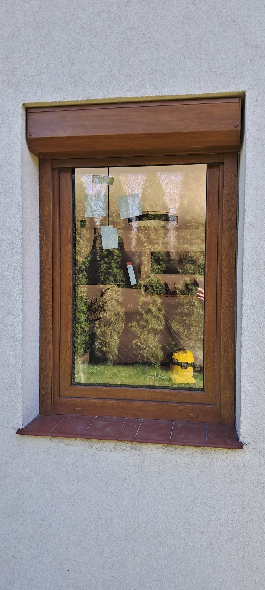 Okno PCV z brązową ramą i roletą zewnętrzną zwiniętą u góry, widok na ogród z naklejonymi na szybie kartkami i stojącym na parapecie od wewnątrz żółtym odkurzaczem.