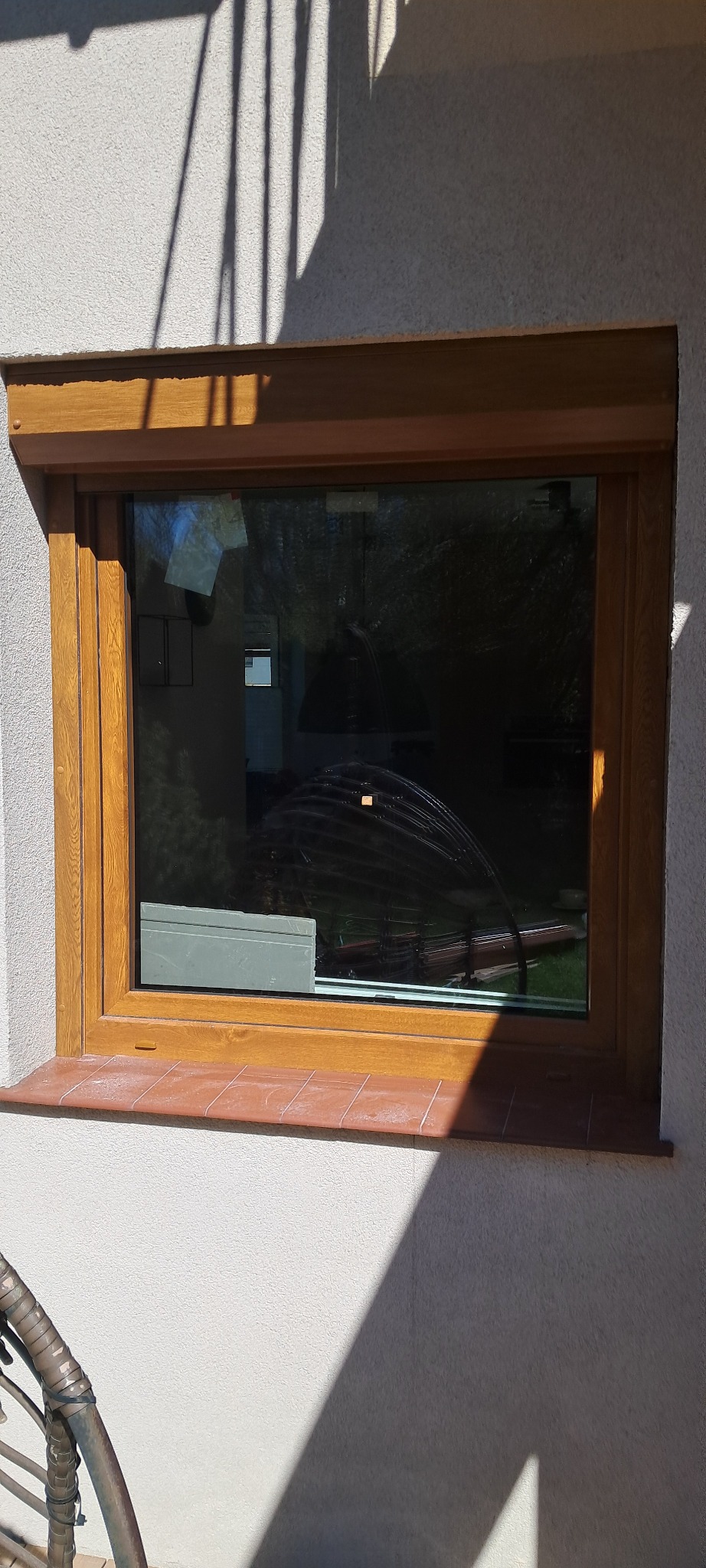 Brązowa rama okienna z zamontowaną roletą zewnętrzną, widoczna z zewnątrz budynku, z terakotowym parapetem i odbiciem w szybie.