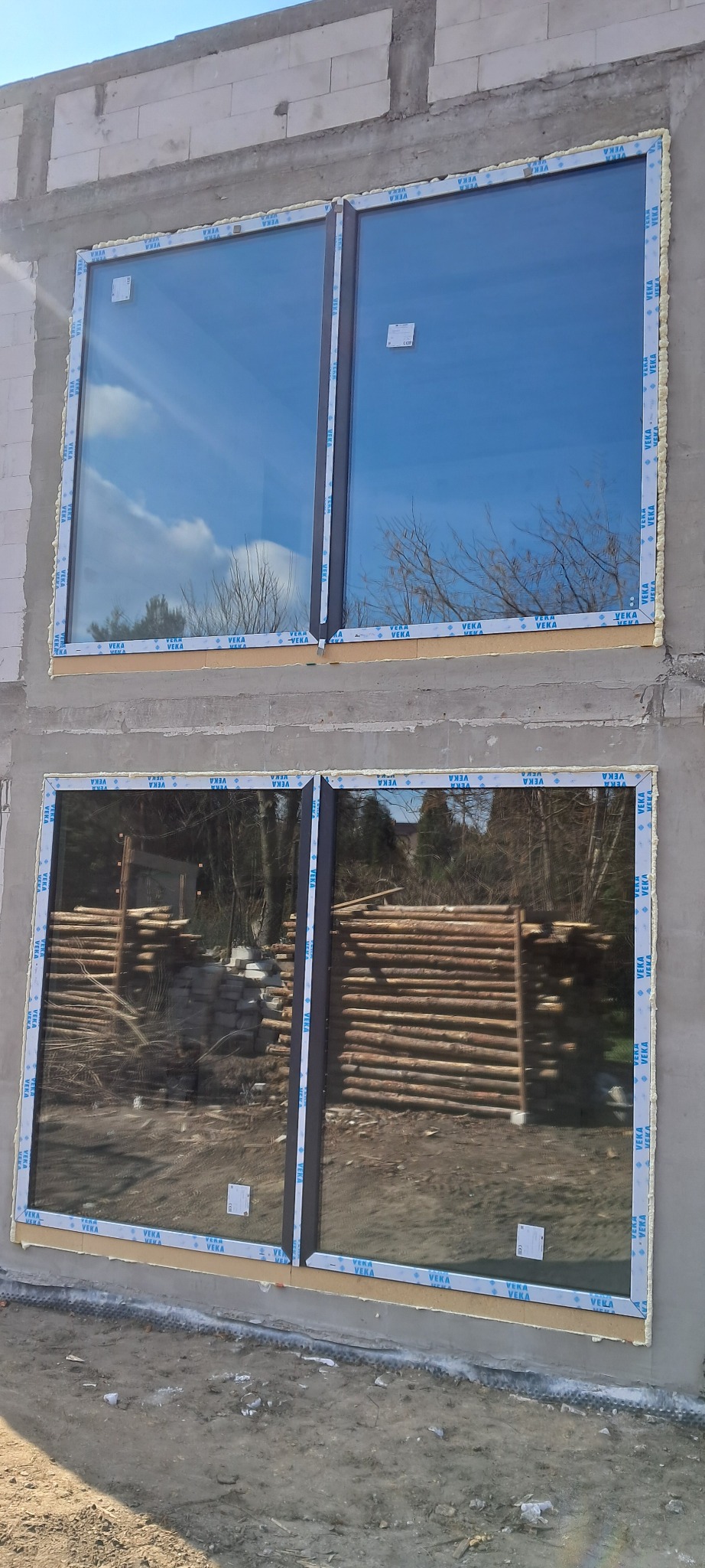 Dwa okna zamontowane w surowym murze, zabezpieczone folią ochronną z logo producenta, pianka montażowa widoczna w szczelinach, w górnym oknie odbija się błękitne niebo, w dolnym stos drewna.
