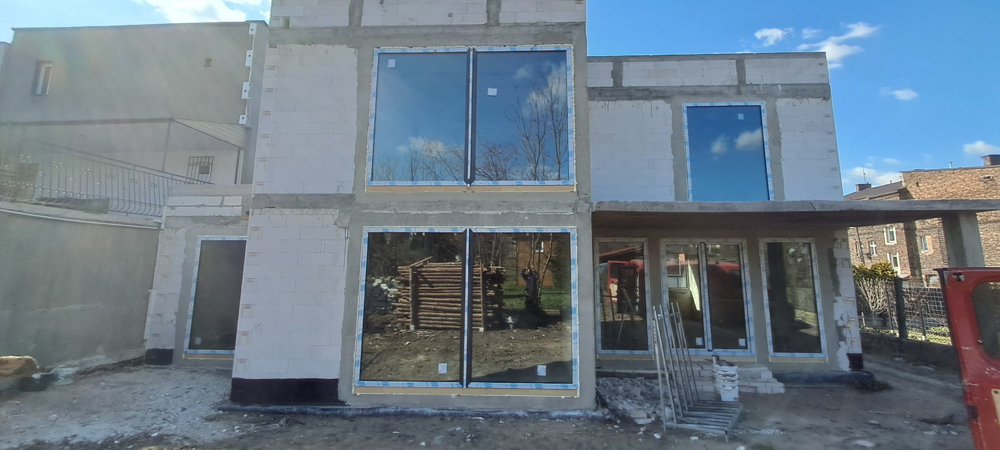 Surowy stan domu w trakcie budowy z zamontowanymi dużymi oknami z wyraźnym odbiciem nieba i otoczenia, widoczne pustaki i brak wykończenia elewacji.