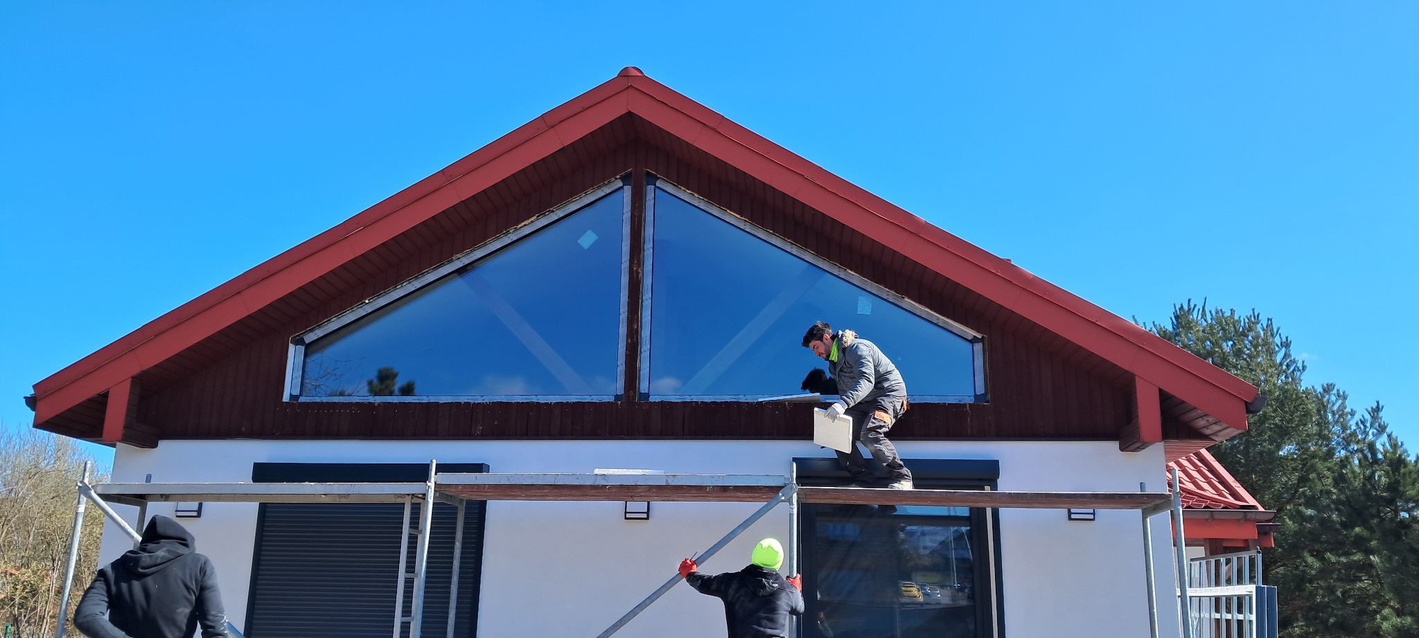 Montażysta na rusztowaniu, instalujący okno trójkątne pod dachem domu z czerwonymi elementami drewnianymi, widoczne również rolety zewnętrzne na niższych oknach.