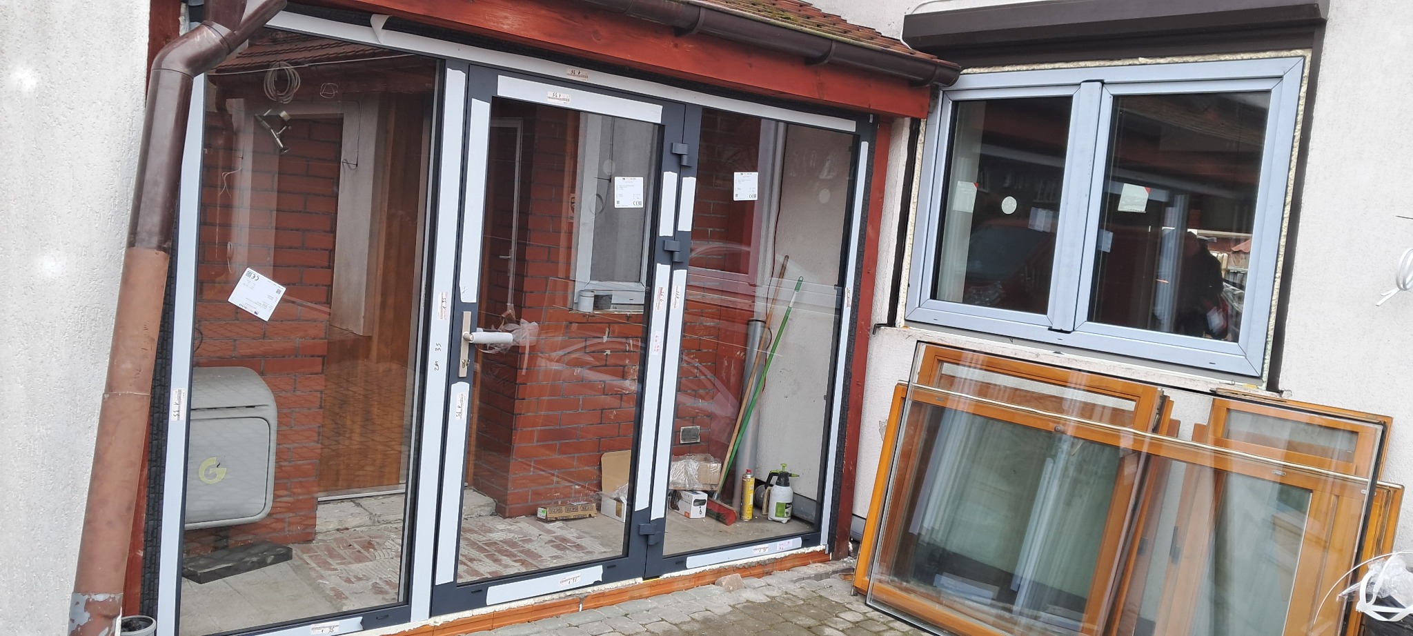 Montaż szklanych drzwi przesuwnych z szarymi ramami; obok okno z białą ramą i stos drewnianych ram okiennych; widoczne narzędzia i akcesoria montażowe.