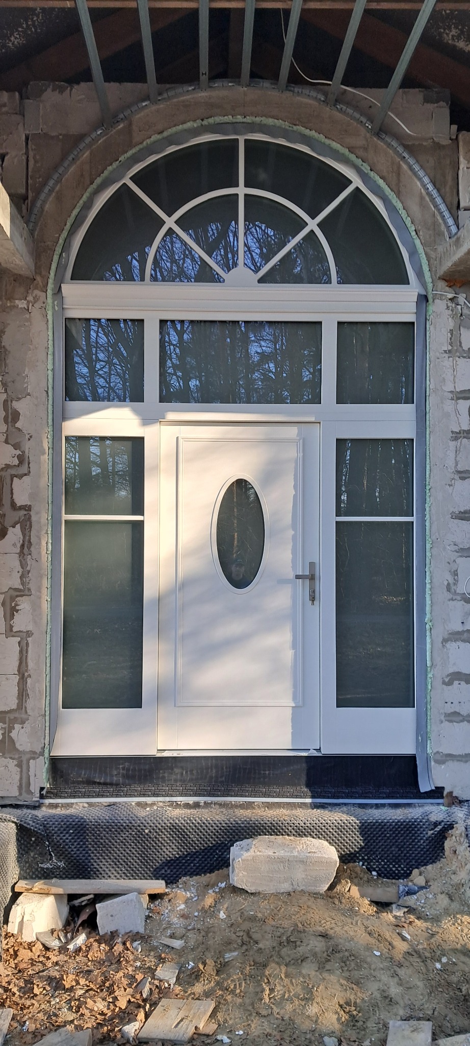 Białe drzwi wejściowe z owalnym przeszkleniem i łukowym naświetlem, zamontowane w surowym stanie budynku w Będzinie, widoczne pianka montażowa i niezakończone prace wykończeniowe.