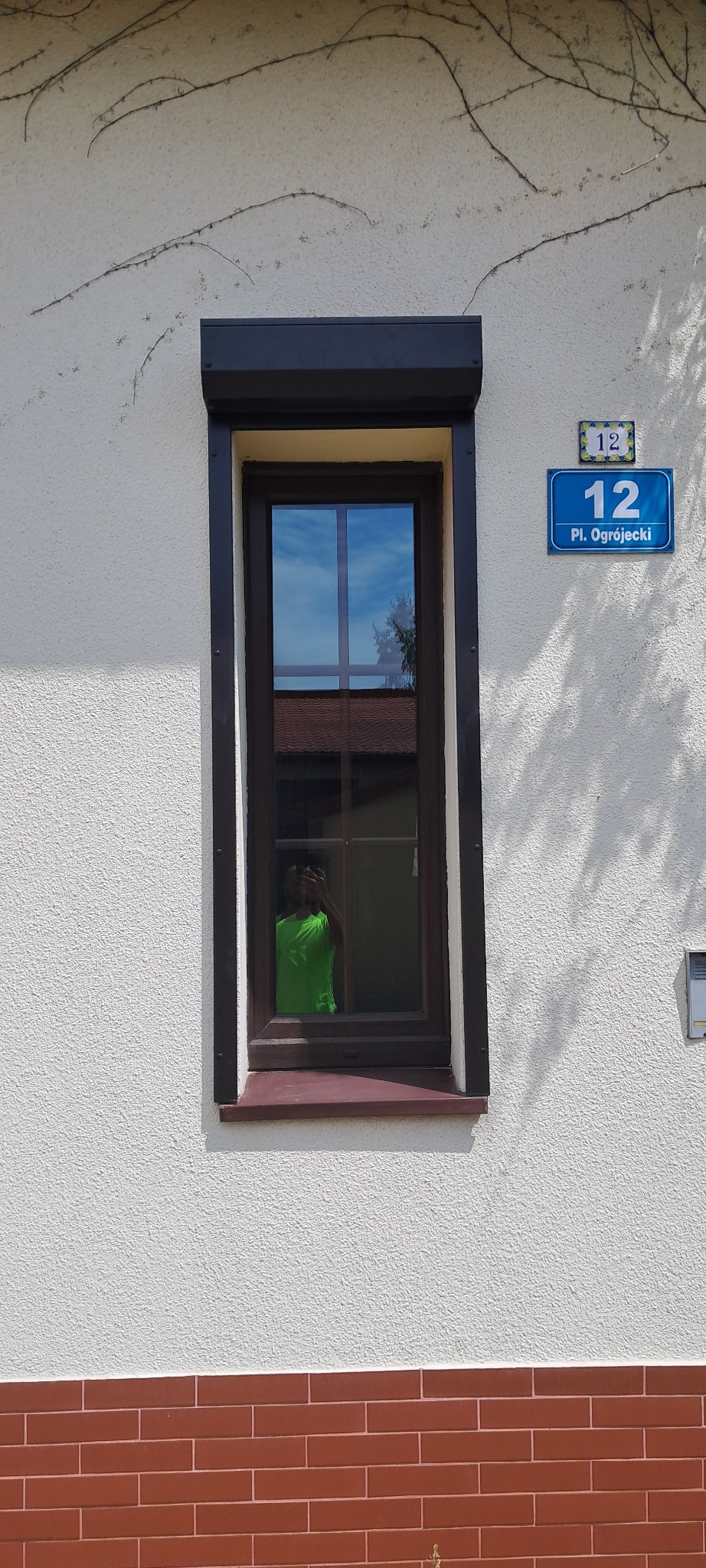 Zamknięte okno PCV z brązową ramą i roletą zewnętrzną na elewacji domu z numerem 12, odbicie osoby w zielonej koszulce w szybie.