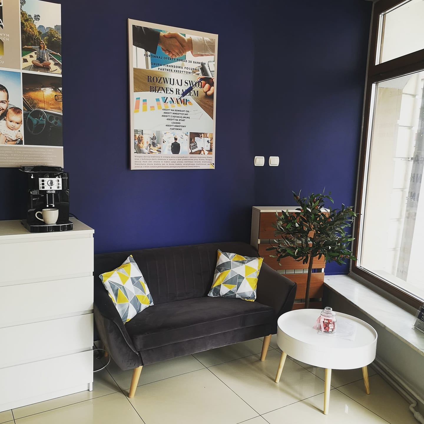 Poczekalnia biura z granatową ścianą, na której wisi plakat reklamujący usługi finansowe, szara sofa z geometrycznymi poduszkami, biały stolik kawowy z dekoracją, ekspres do kawy na komodzie...