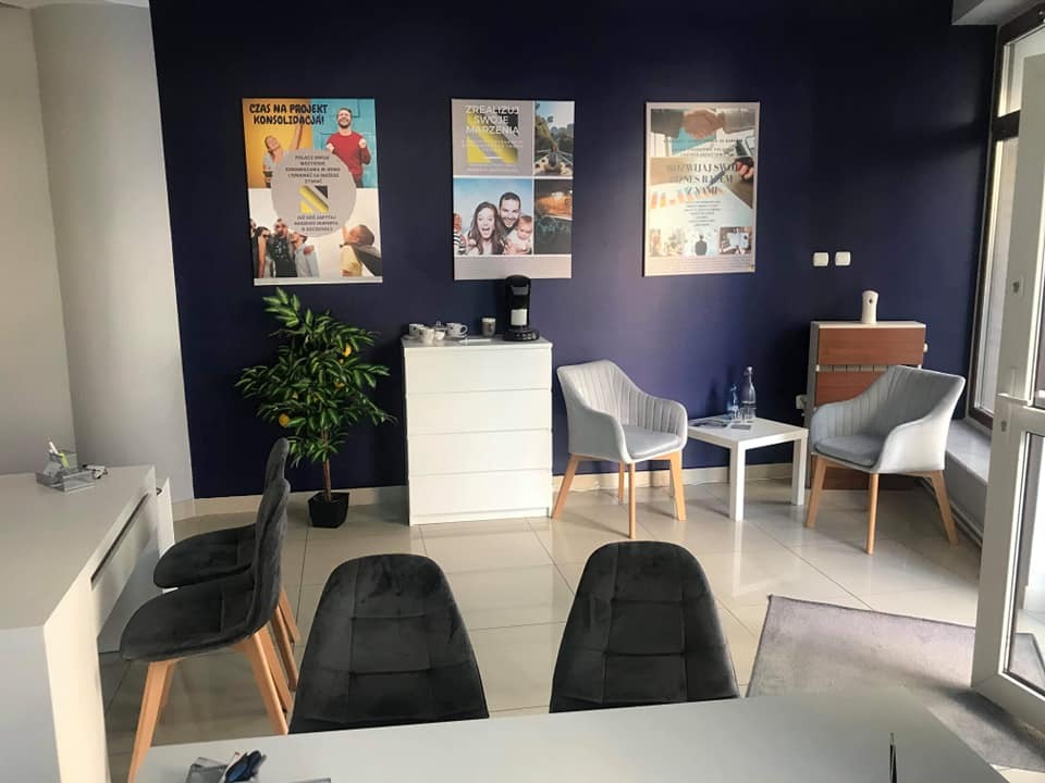 Jasne biuro z ciemnoniebieską ścianą, plakatami i minimalistycznymi meblami, w tle mała roślina w doniczce i ekspres do kawy na białej komodzie.