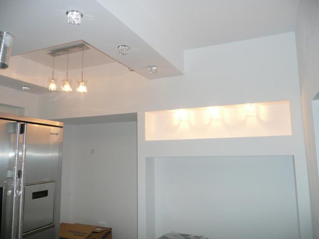 Białe wnętrze z oświetleniem: sufit podwieszany z halogenami i lampami wiszącymi nad chłodziarko-zamrażarką, wnęka ścienna z podświetleniem, kartony na podłodze.