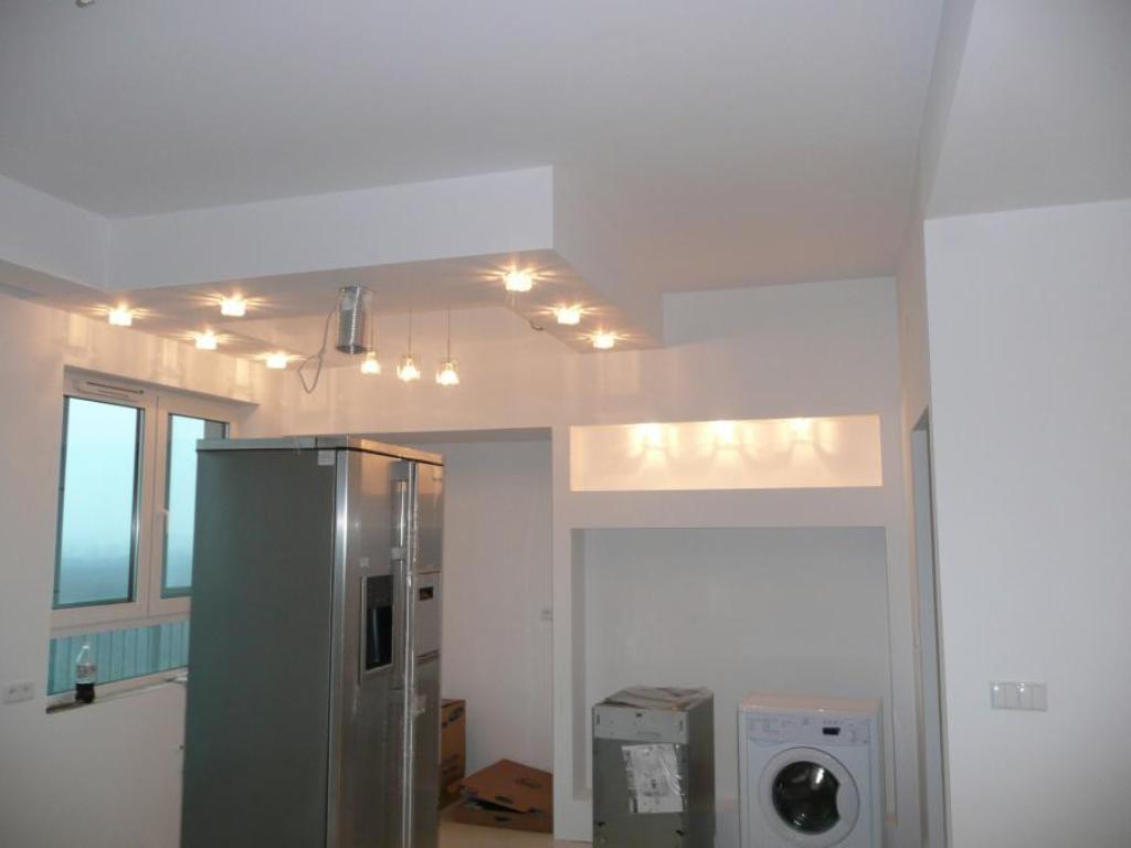 Nowoczesny sufit podwieszany z oświetleniem punktowym w kuchni, widoczne ścianki działowe, lodówka i pralka w trakcie remontu.