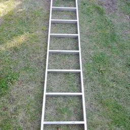 Aluminiowa drabina przystawna leżąca na trawie, widok z góry, siedem szczebli, cień na trawie.