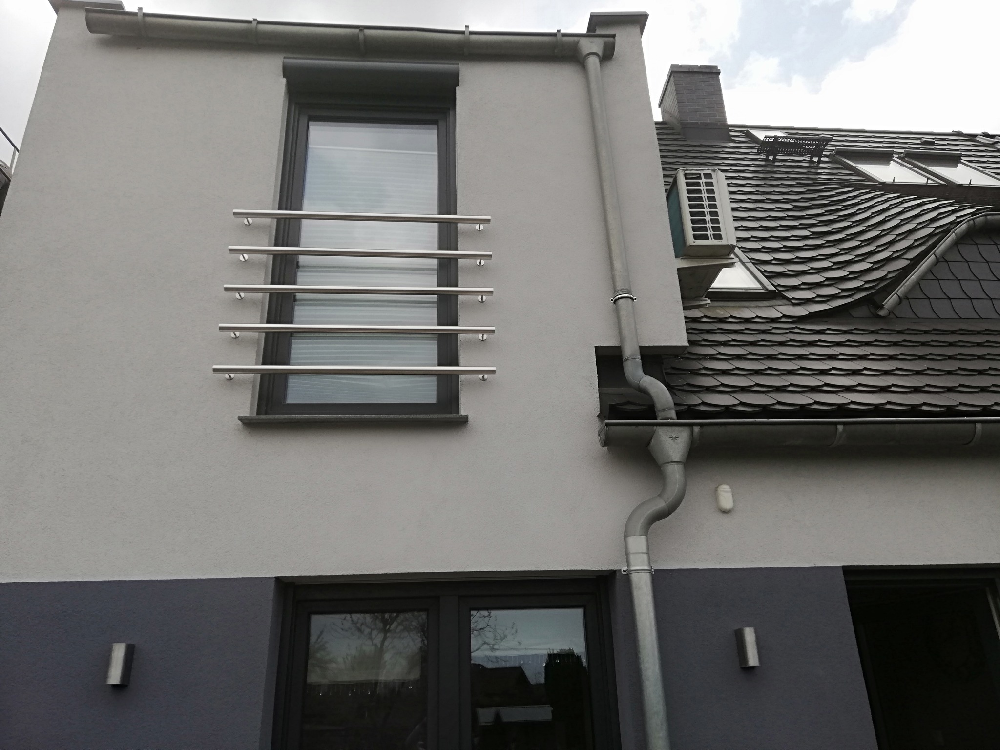 Elewacja budynku z oknem zabezpieczonym poziomymi, metalowymi prętami, widoczny system rynnowy i fragment dachu z oknami połaciowymi.
