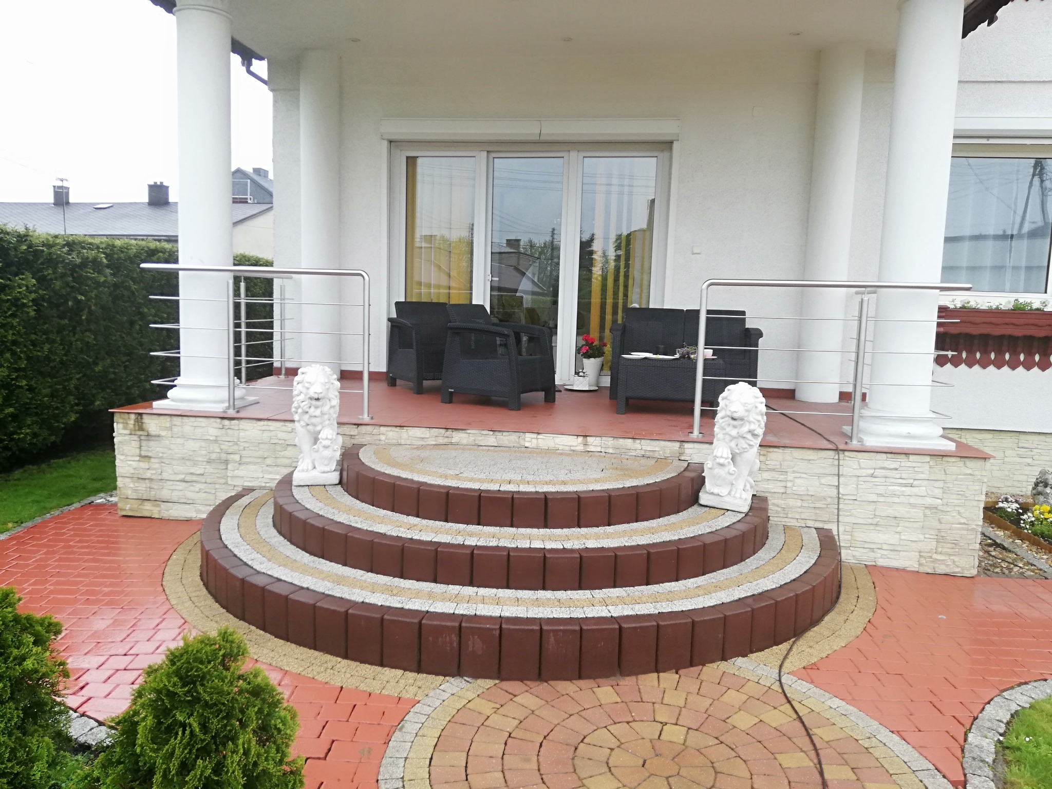 Zewnętrzne schody z brązowej kostki brukowej z białymi akcentami, prowadzące na taras z meblami ogrodowymi i balustradą ze stali nierdzewnej, ozdobione rzeźbami lwów.