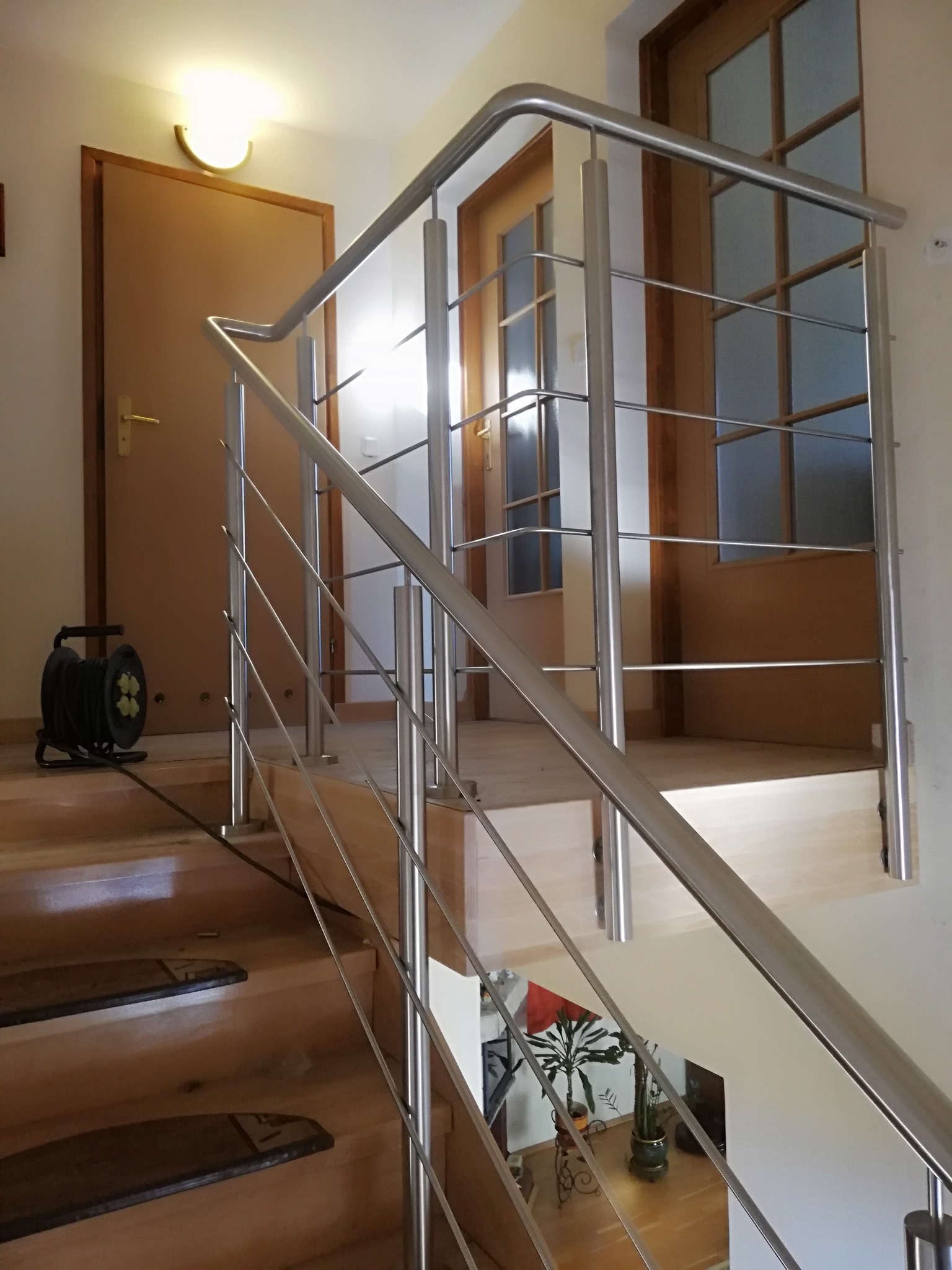 Schody z drewnianymi stopniami i nowoczesną balustradą ze stali nierdzewnej; w tle widoczne drzwi i zwinięty przedłużacz elektryczny.