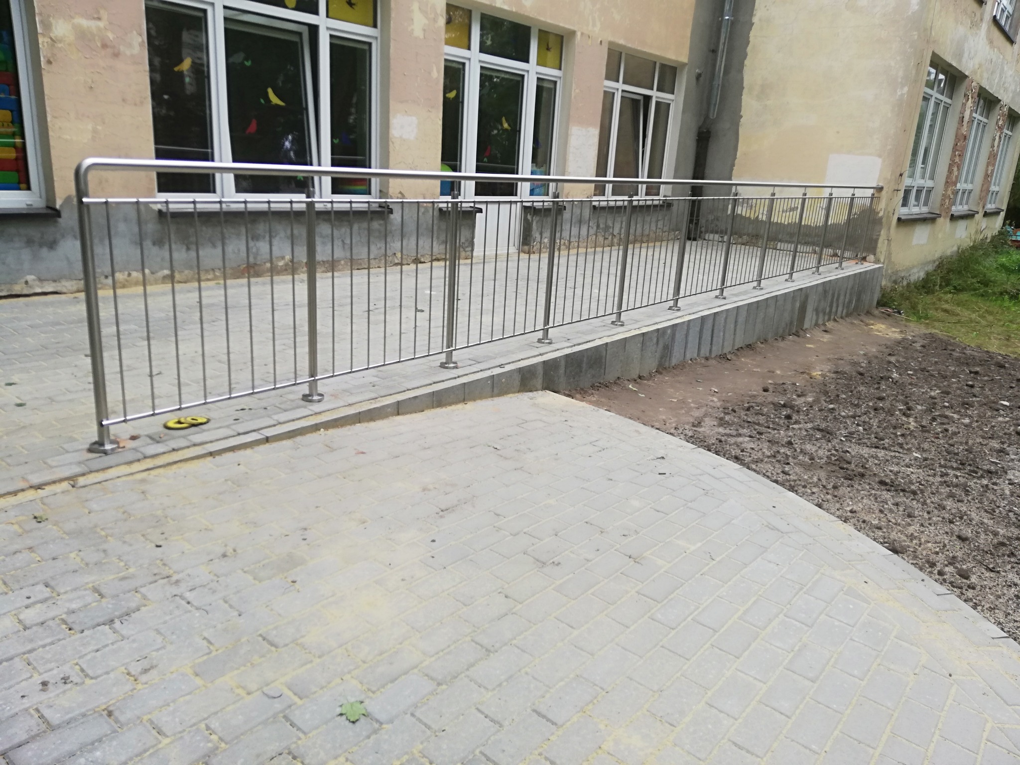 Zewnętrzna balustrada z nierdzewnej stali przed budynkiem z oknami, zamontowana na podwyższeniu z kostki brukowej, widoczne niedokończone prace ziemne obok.