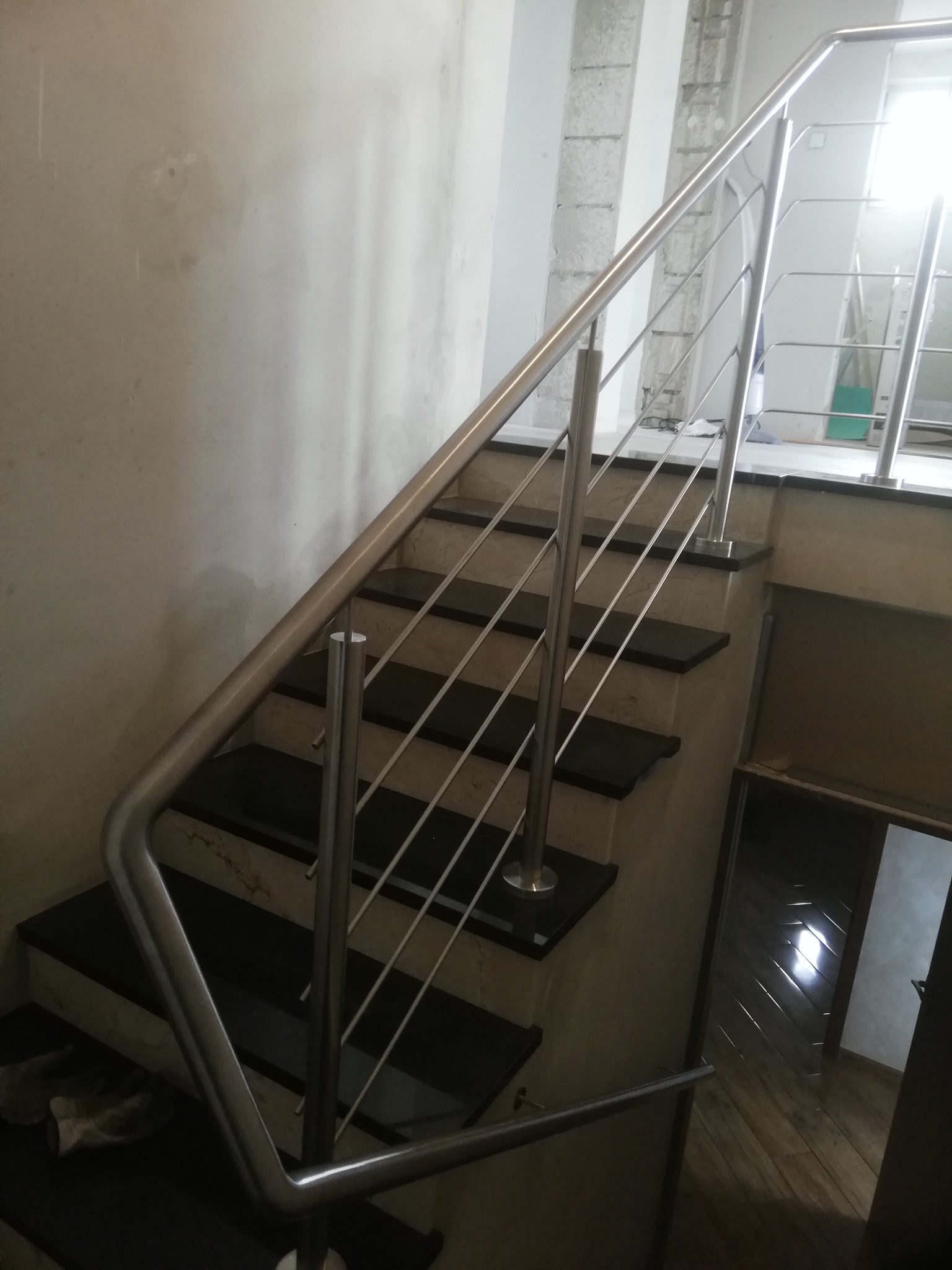 Schody z czarnymi stopniami i balustradą ze stali nierdzewnej, widoczne ślady prac wykończeniowych na ścianach.