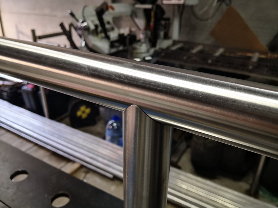 Szczegółowe ujęcie spawu kątowego na nierdzewnej poręczy, widoczne ślady obróbki i maszyna do cięcia w tle.