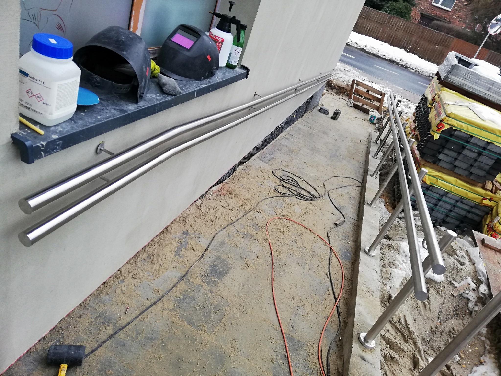 Montaż dwupoziomowej, metalowej poręczy przy schodach zewnętrznych, widoczne narzędzia i materiały budowlane, w tle ulica i resztki śniegu.