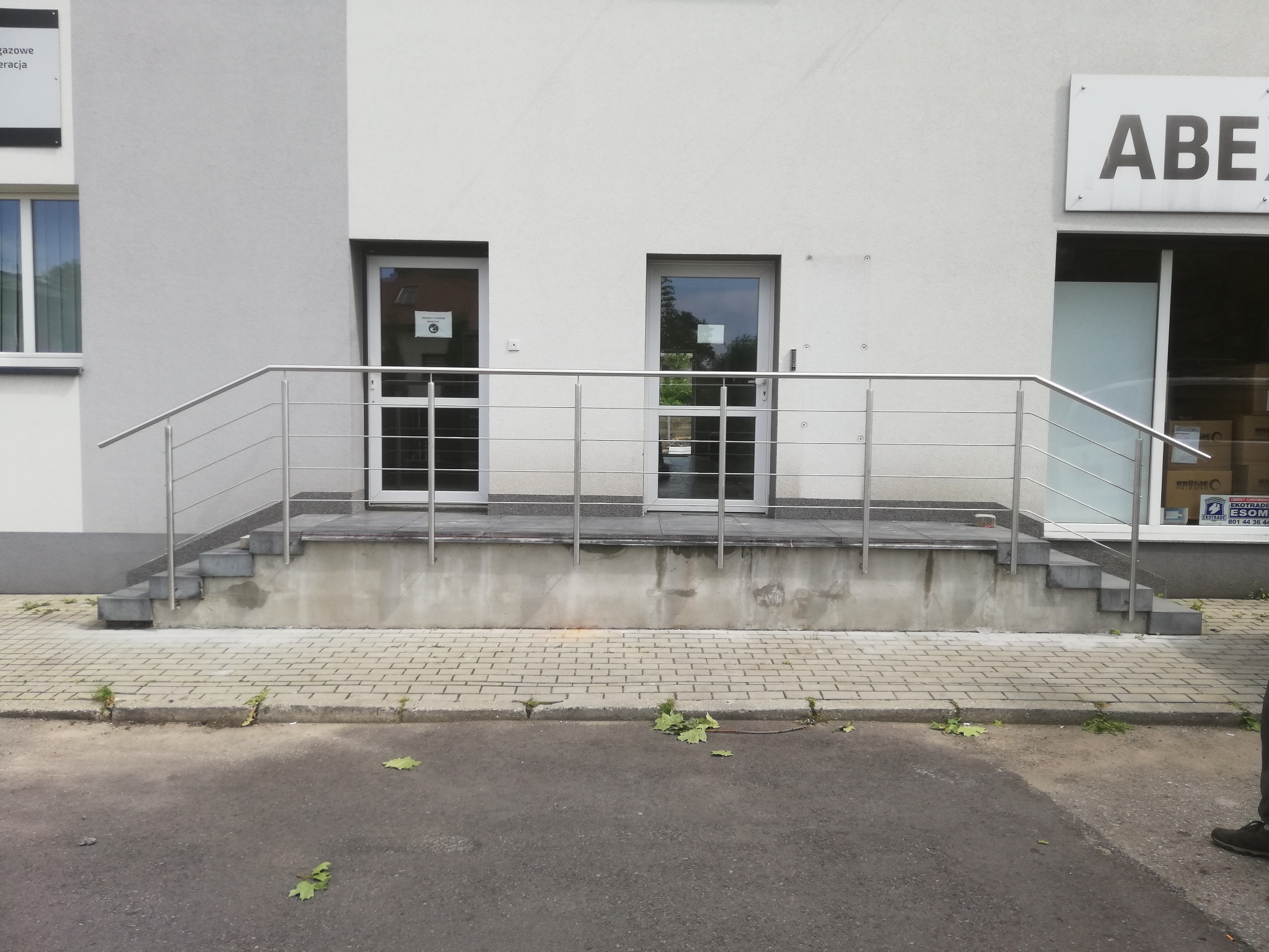Zewnętrzne schody wejściowe do budynku z dwiema parami drzwi zabezpieczone metalową, prostą barierką ze stali nierdzewnej.