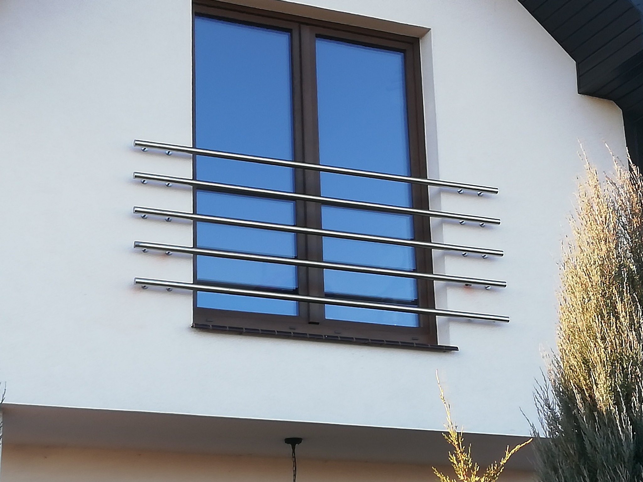 Zewnętrzna, pozioma, metalowa barierka okienna z rurek, zamontowana na białej elewacji domu, z widocznym oknem z brązową ramą i odbiciem nieba w szybie.