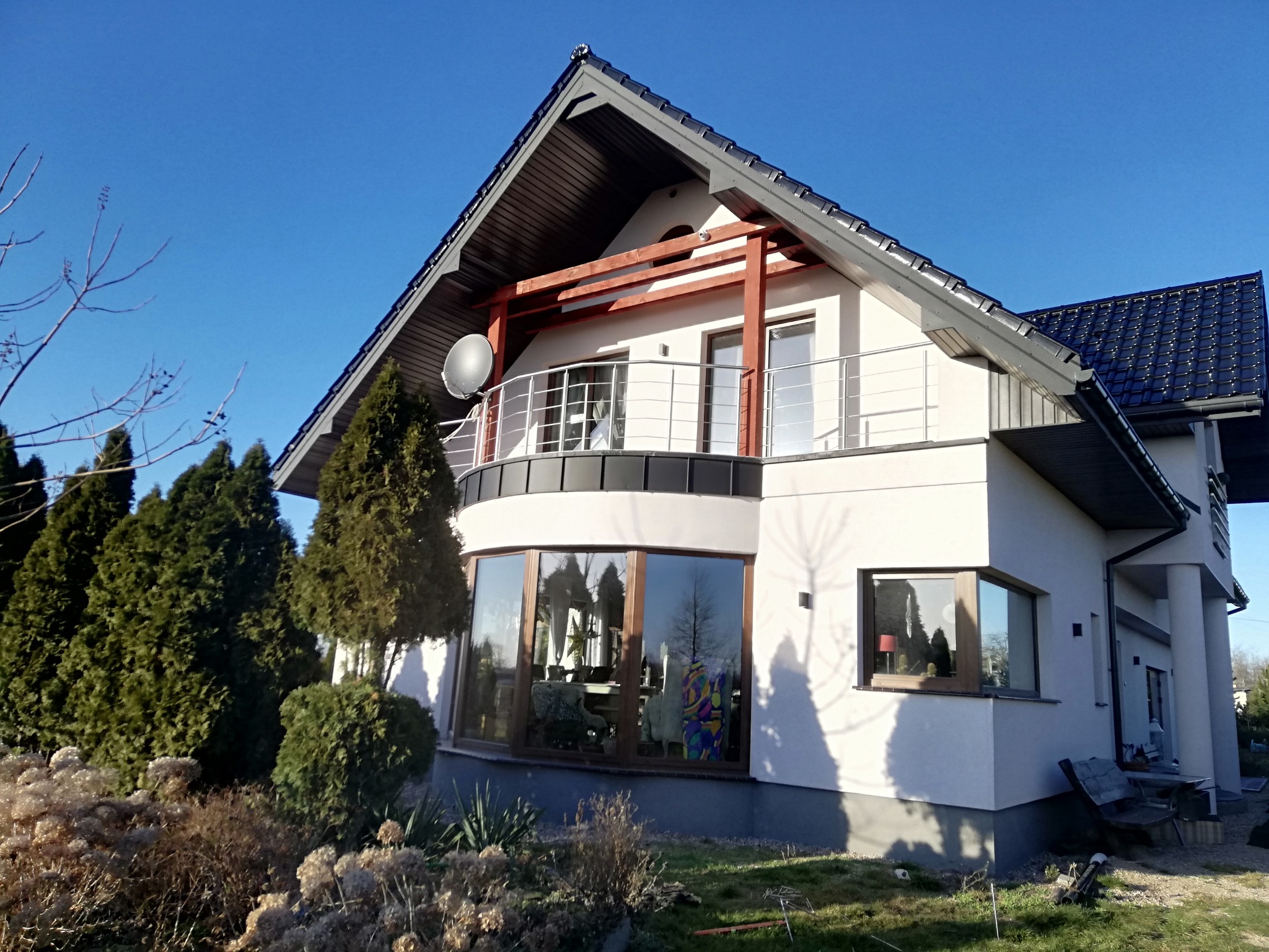 Nowoczesny dom z białą elewacją, balkonem z nierdzewną balustradą, drewnianą pergolą i dużym oknem panoramicznym, otoczony zielenią w słoneczny dzień.