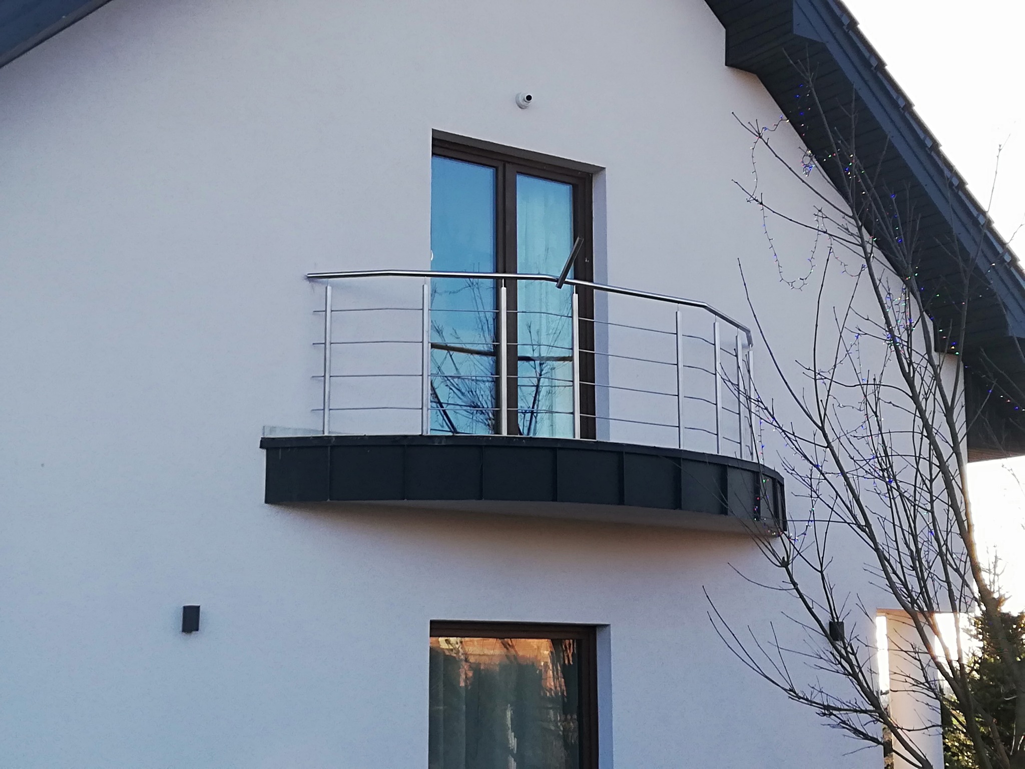 Nierdzewna balustrada balkonowa z poziomymi prętami, zamontowana na zaokrąglonym balkonie nowoczesnego domu z białymi ścianami, widoczny fragment drzewa z lampkami.
