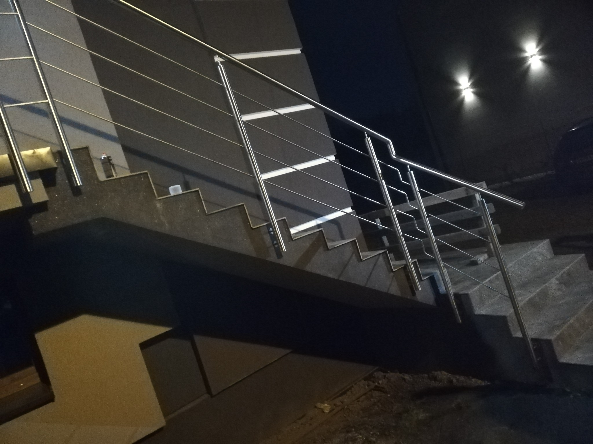 Zewnętrzne schody z ciemnego kamienia z nowoczesną, minimalistyczną balustradą ze stali nierdzewnej, oświetlone przez dwa reflektory na ścianie budynku.