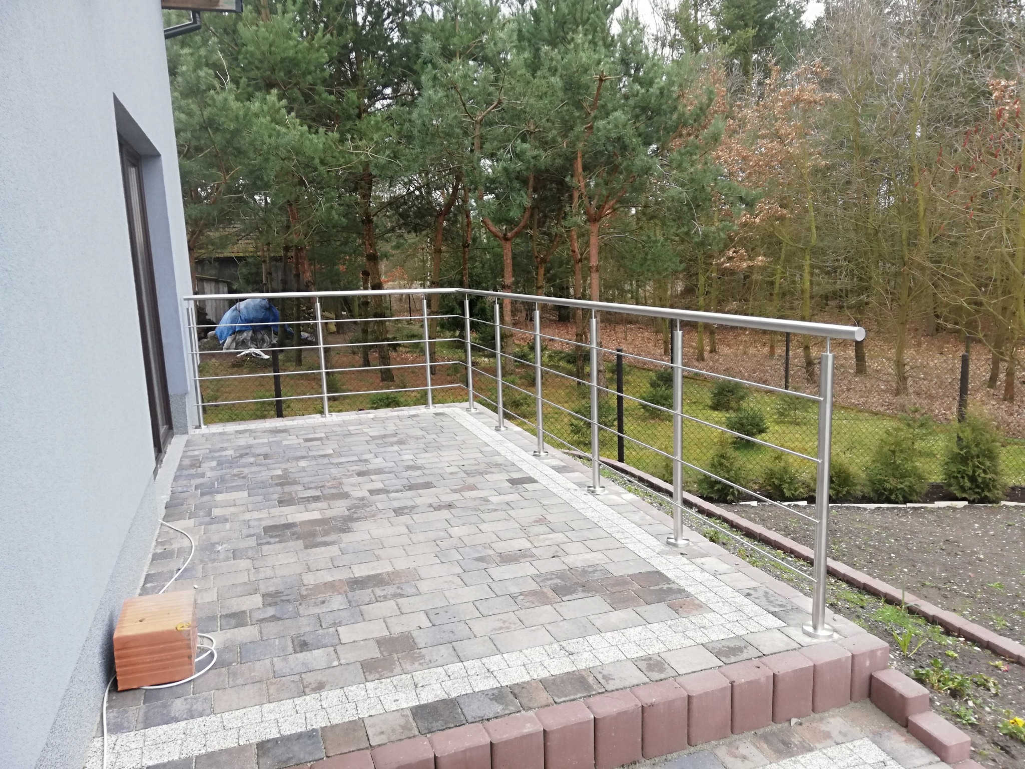Balkon z nierdzewną balustradą, widok na las, kostka brukowa na podłodze, pudełko przy ścianie.