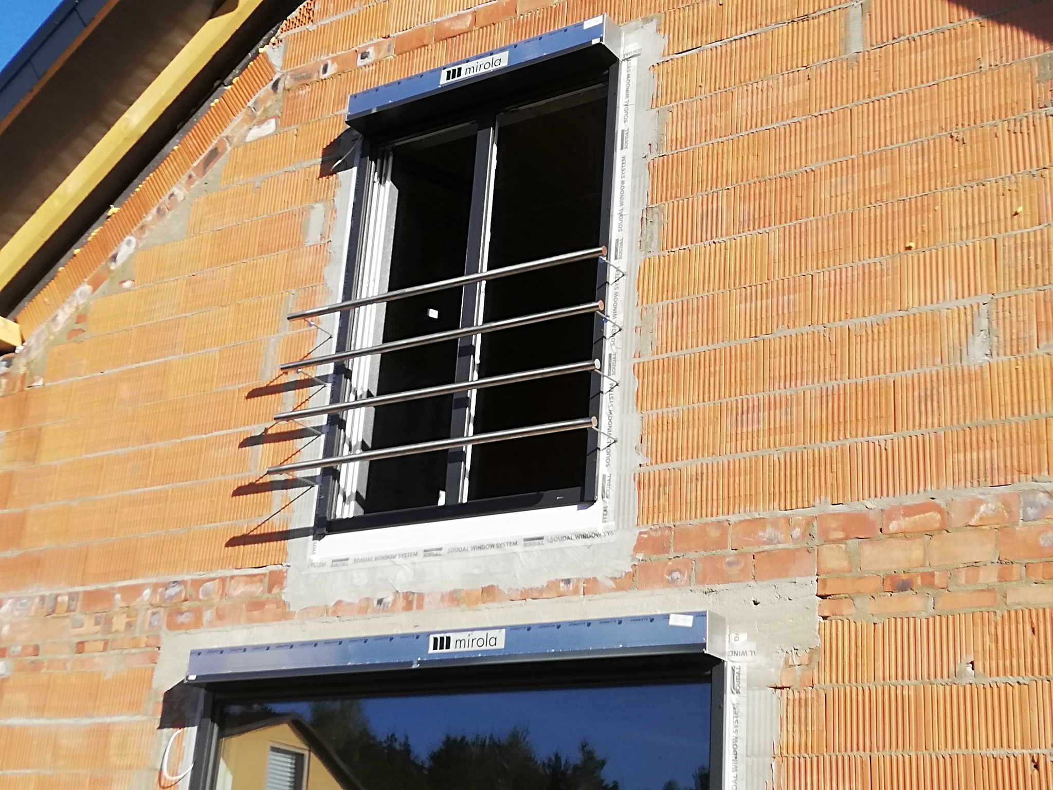 Montaż zabezpieczenia okiennego z poziomych, metalowych prętów na surowej ścianie z czerwonej cegły porotherm. Widoczne okna z roletami zewnętrznymi i logo producenta stolarki okiennej.