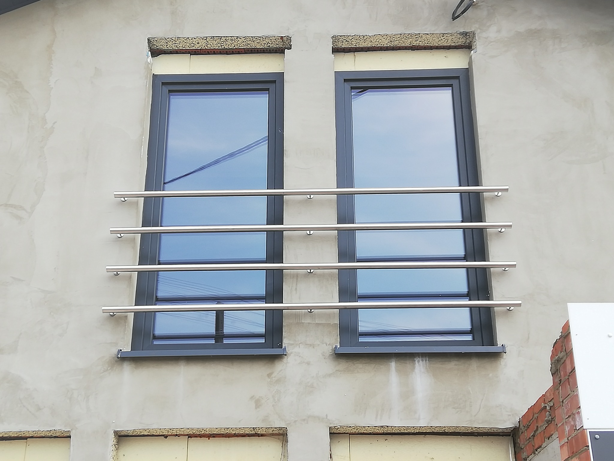 Dwa okna z szarymi ramami zabezpieczone poziomymi, metalowymi rurkami na surowej elewacji budynku w trakcie wykończenia.