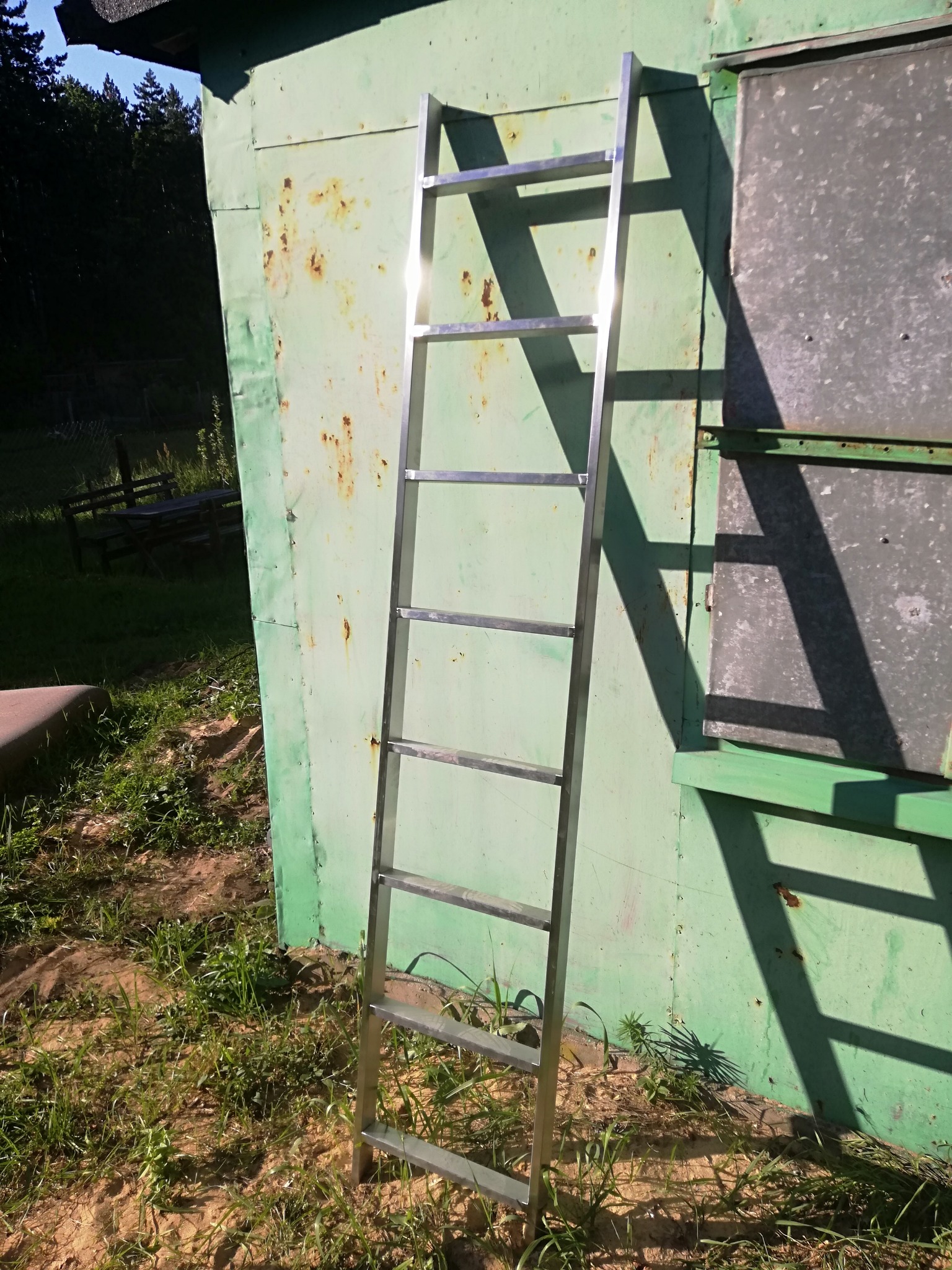 Aluminiowa drabina przystawna oparta o pomalowaną na zielono, blaszana ścianę budynku gospodarczego z widocznymi śladami korozji.