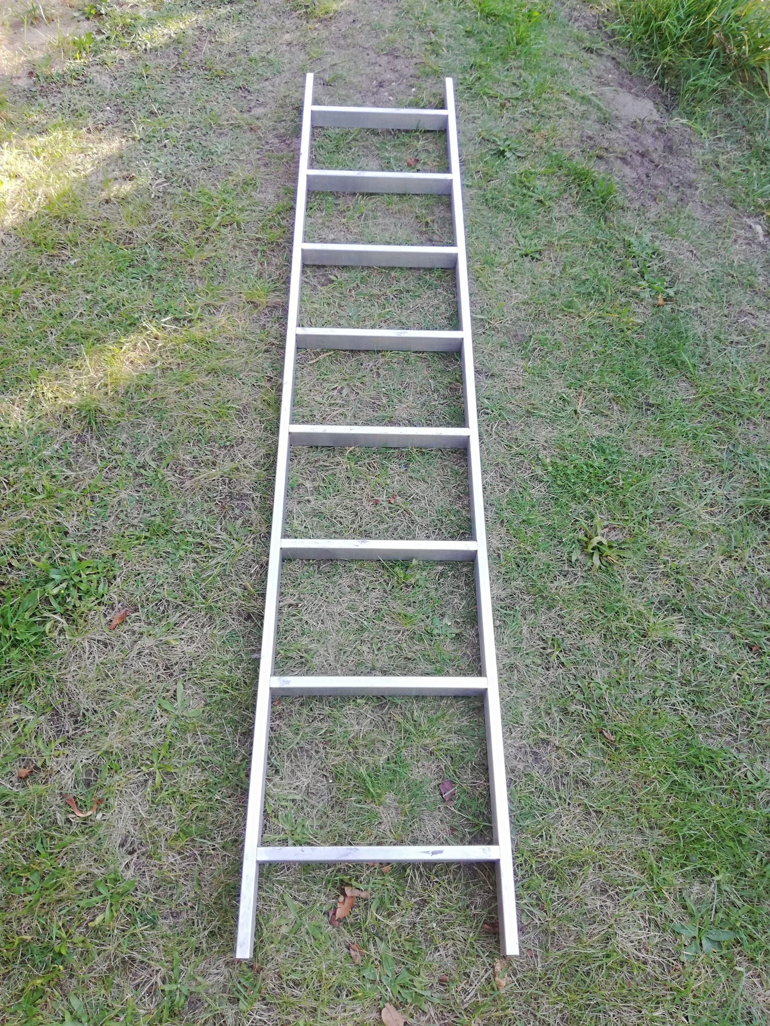 Aluminiowa drabina przystawna leżąca na trawie, widok z góry, siedem szczebli, cień na trawie.