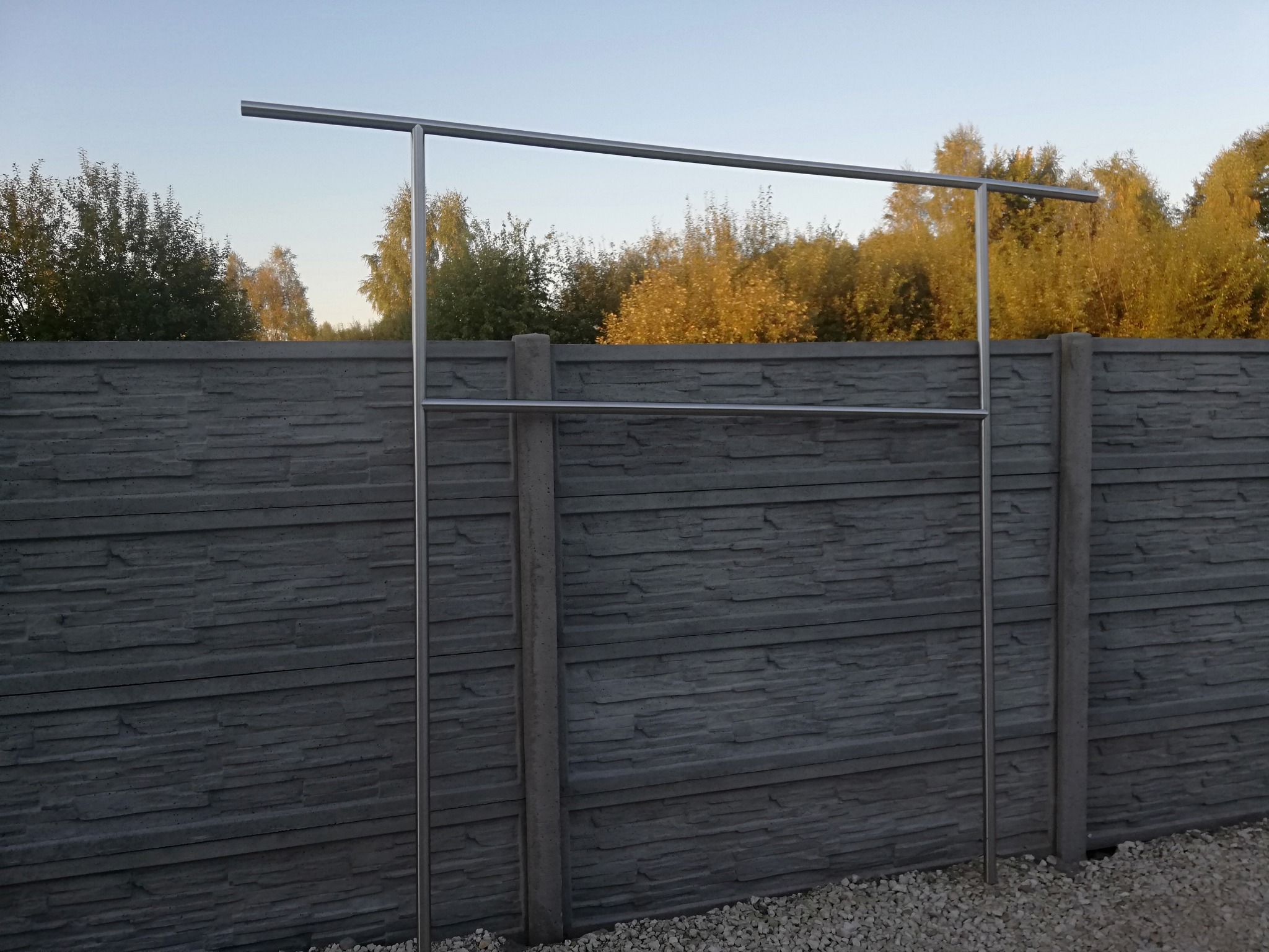 Stalowa konstrukcja z poziomymi poprzeczkami, prawdopodobnie przygotowana do montażu szkła, zamocowana przed betonowym ogrodzeniem panelowym.