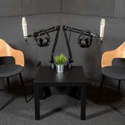 Dwa mikrofony studyjne z słuchawkami na wysięgnikach, ustawione symetrycznie po obu stronach małego czarnego stolika z doniczką z zieloną rośliną, pomiędzy dwoma drewnianymi krzesłami na tle...