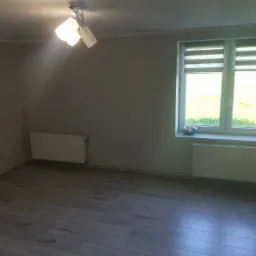 Pusty pokój z szarymi ścianami, panelami podłogowymi imitującymi drewno, białym grzejnikiem i oknem z roletami dzień-noc.