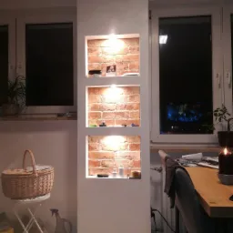 Wnęka ścienna z cegły, podświetlona trzema lampkami, z dekoracjami i przedmiotami na półkach, w tle okno z widokiem na nocne miasto.
