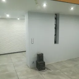 Wnętrze biura po odświeżeniu: ściany pomalowane na szaro, biała cegła dekoracyjna, szare płytki podłogowe, widoczny fragment instalacji elektrycznej i starego sprzętu audio.