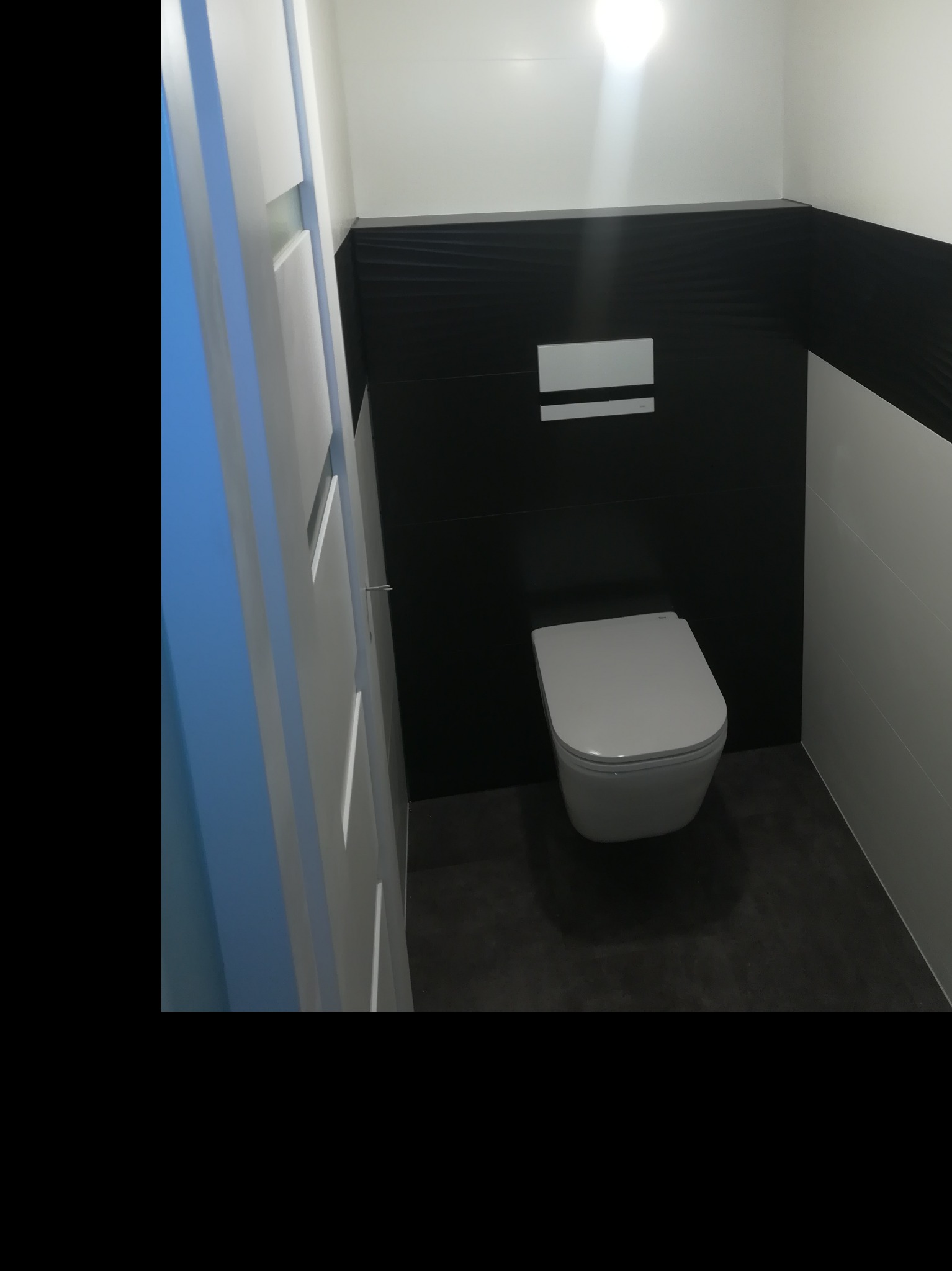 Nowoczesna toaleta podwieszana w łazience z czarno-białymi płytkami, widok na przycisk spłukujący i fragment białych drzwi.