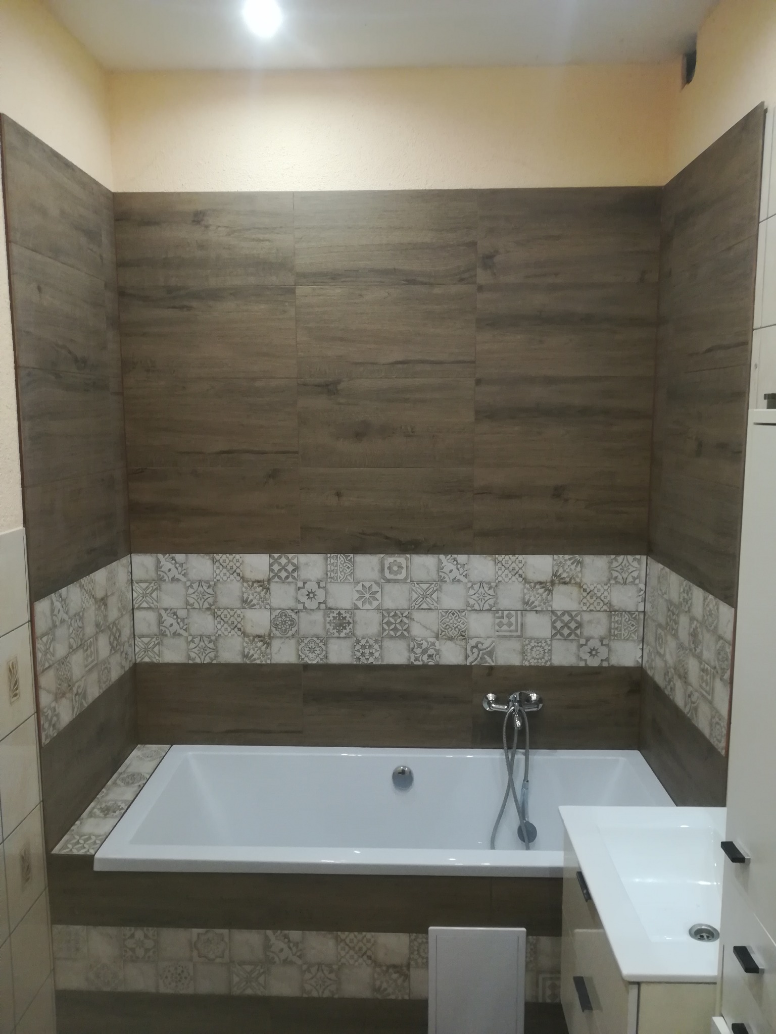 Nowoczesna łazienka z wanną obłożoną brązowymi płytkami imitującymi drewno i dekoracyjnymi kaflami z motywami geometrycznymi, chromowana bateria wannowa, biała umywalka z szafką.