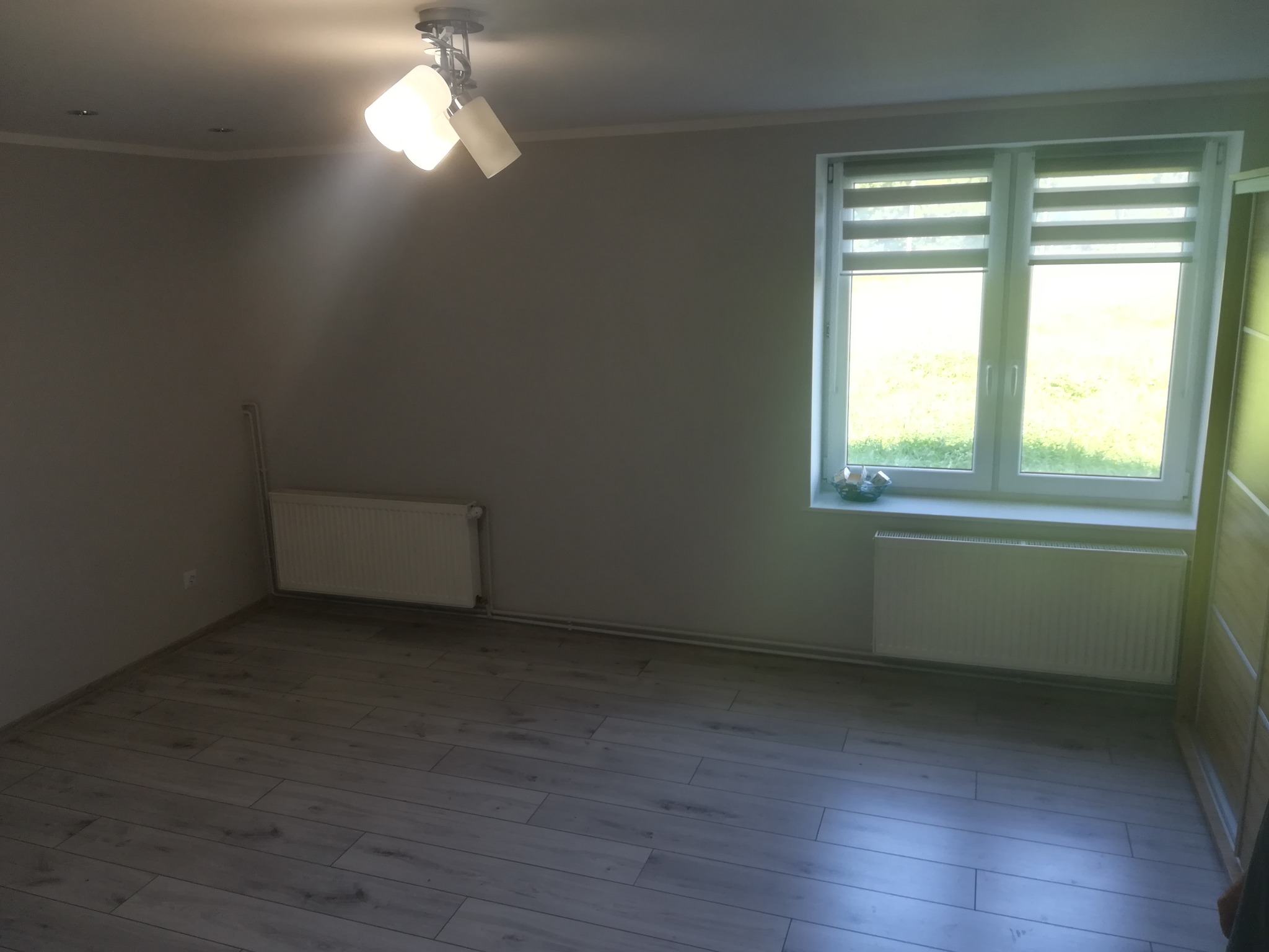 Pusty pokój z szarymi ścianami, panelami podłogowymi imitującymi drewno, białym grzejnikiem i oknem z roletami dzień-noc.