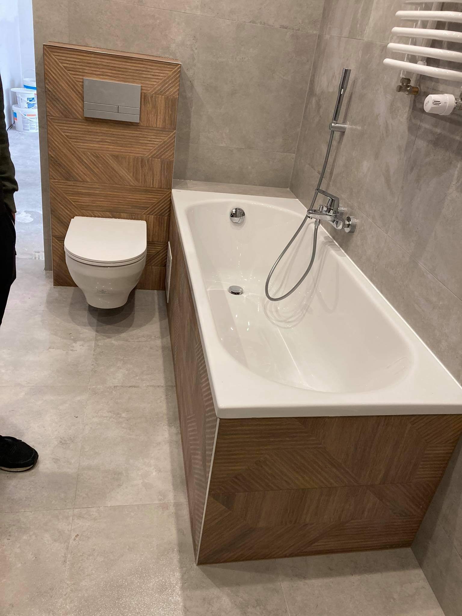 Nowoczesna łazienka z wanną obudowaną drewnopodobnymi panelami, wiszącą toaletą i grzejnikiem drabinkowym na szarej płytce ściennej.